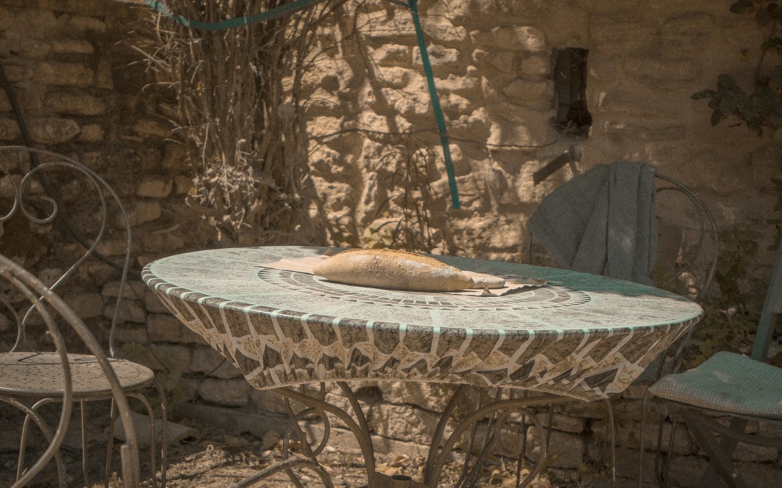 A baguette rests on a mosaic-tiled table beside metal chairs against a stone wall