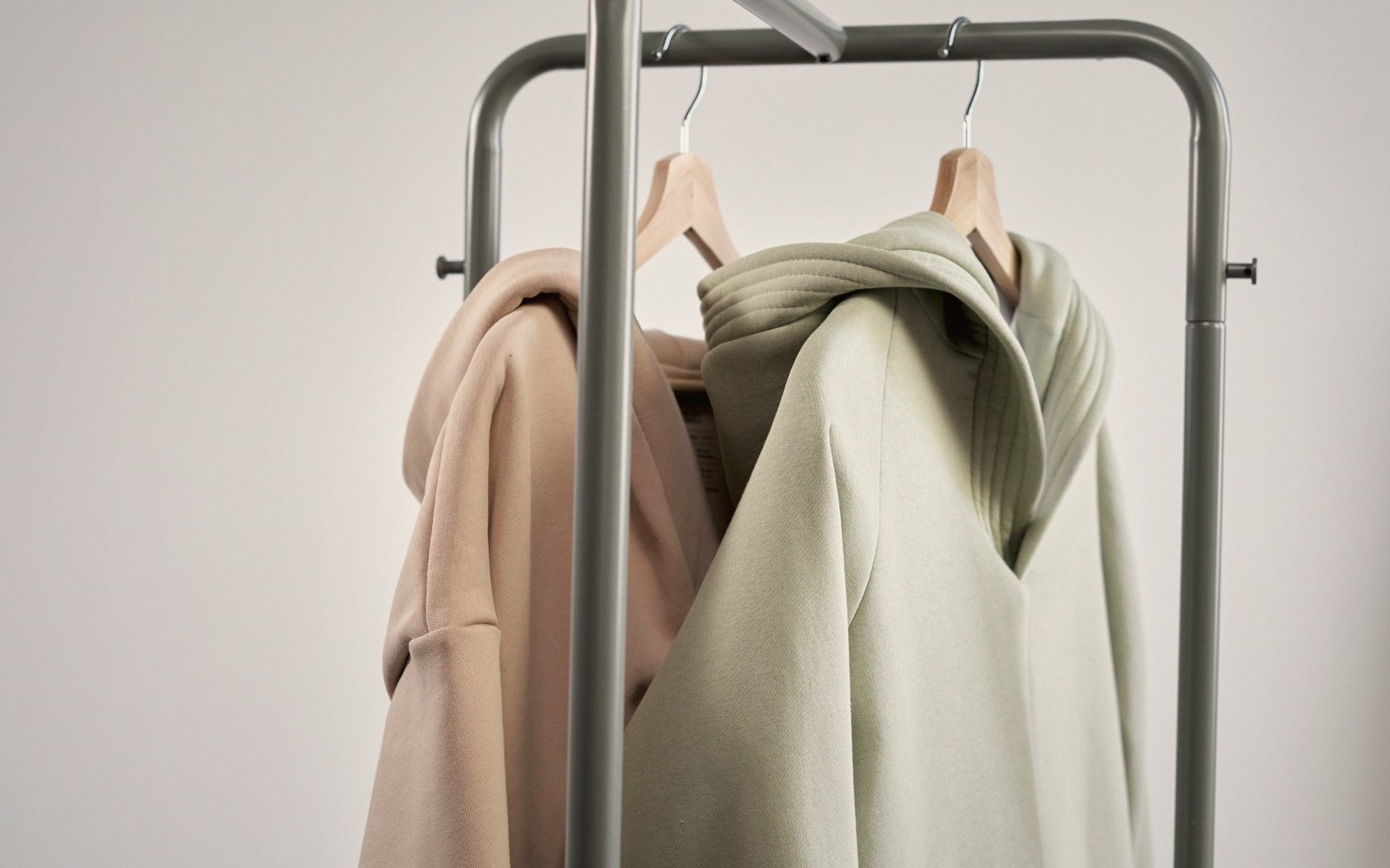 Beige and sage green hoodies hang on wooden hangers from a metal clothing rack