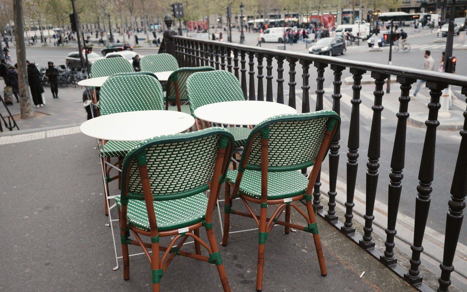 Empty bistro chairs and tables line a railed terrace overlooking a busy city street
